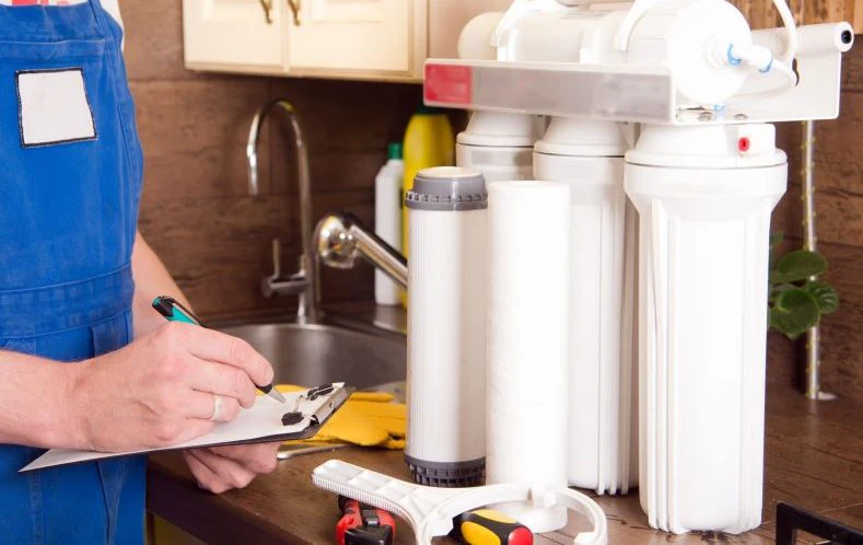 A professional plumber in blue overalls inspecting a multi-stage reverse osmosis water filtration system on a kitchen counter.