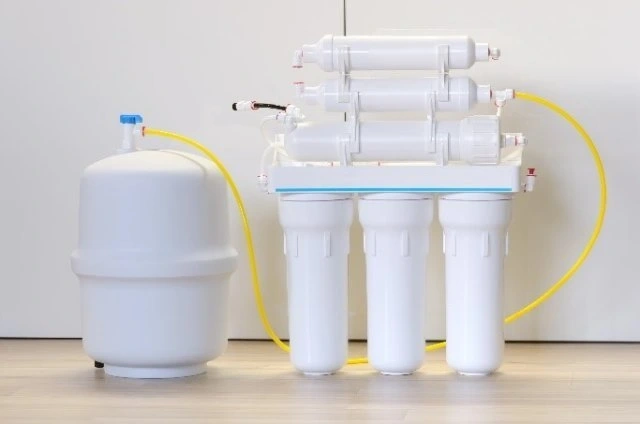 A multi-stage reverse osmosis water filtration system with three vertical pre-filter canisters, three horizontal membrane housings, and a white pressurized storage tank connected by yellow tubing.