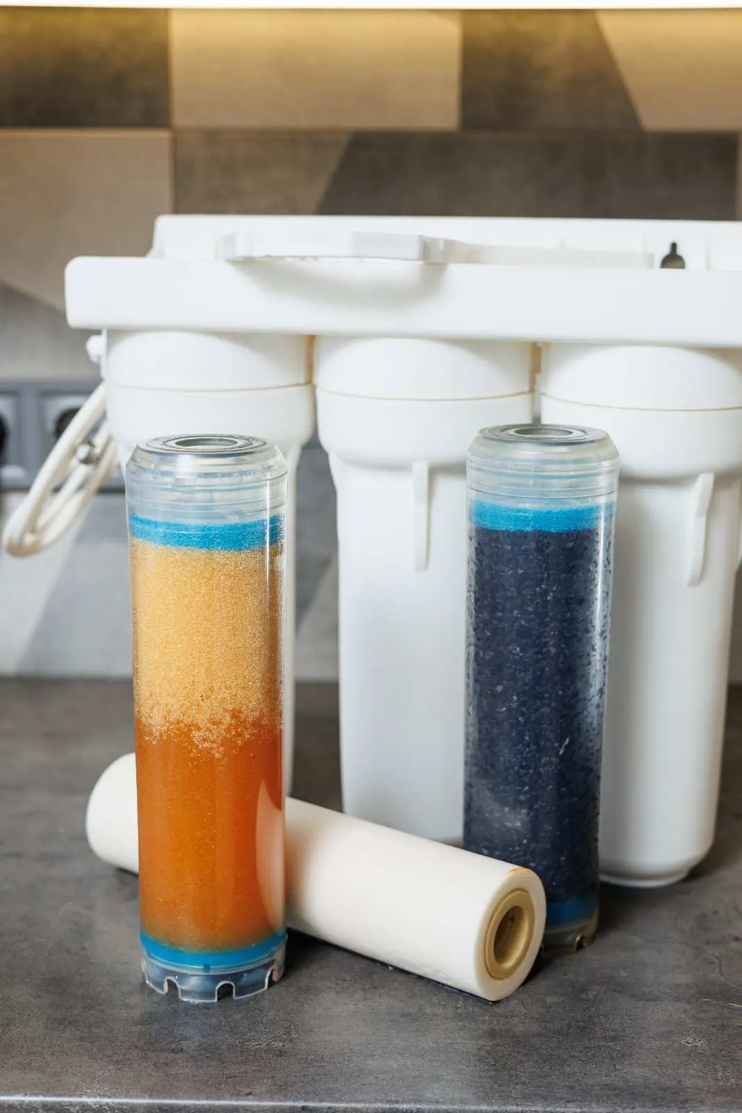 A three-stage under-sink water filtration system with replacement cartridges, including a clear view of an orange sediment filter and a dark activated carbon filter.