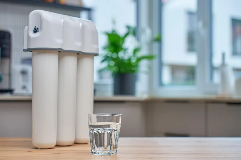 A white three-stage under-sink water filter system standing on a wooden kitchen counter next to a clear glass of fresh water.
