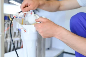 A technician's hands connecting water lines to install a reverse osmosis water filter system under a kitchen sink.