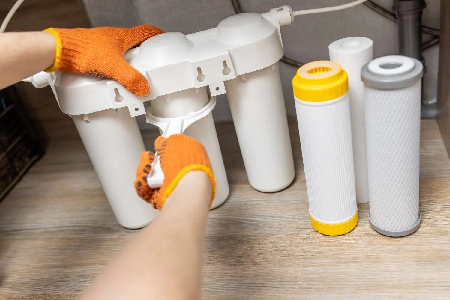 A plumber wearing orange gloves uses a plastic wrench to tighten a filter housing on a three-stage reverse osmosis water filtration system under a kitchen sink.