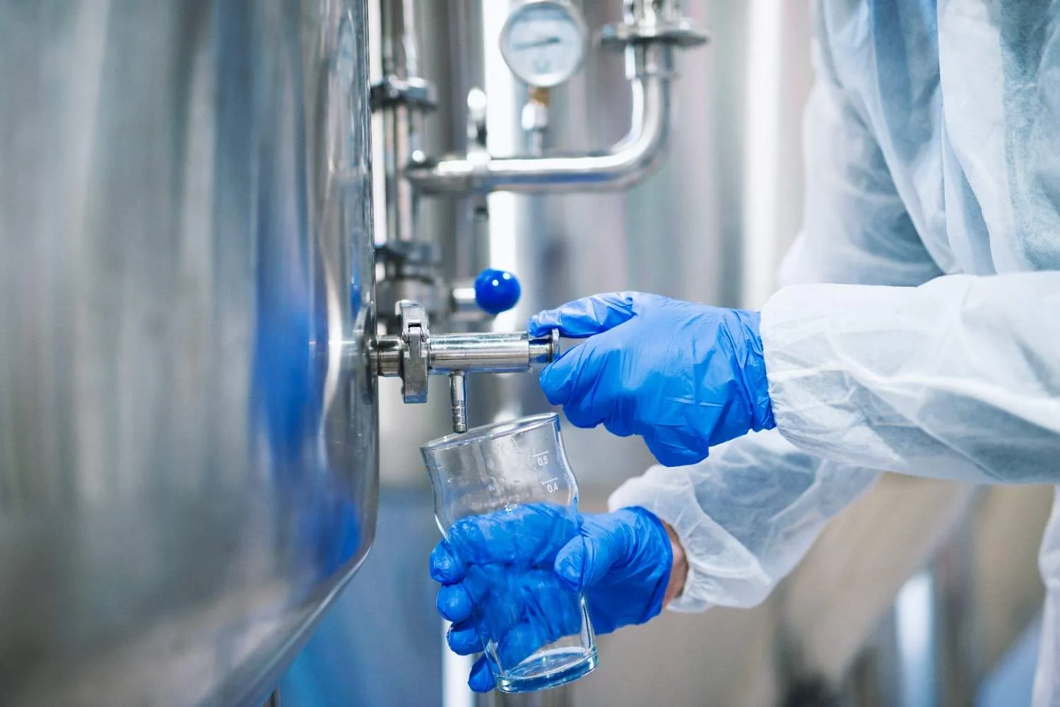 A technician in blue protective gloves and lab gear taking a water sample from a stainless steel industrial tank valve into a glass beaker.