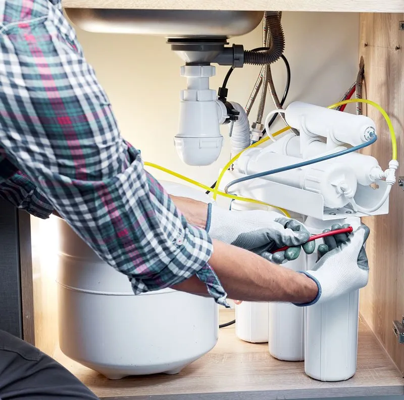 A plumber in a plaid shirt and work gloves installing a multi-stage reverse osmosis water filtration system under a kitchen sink.