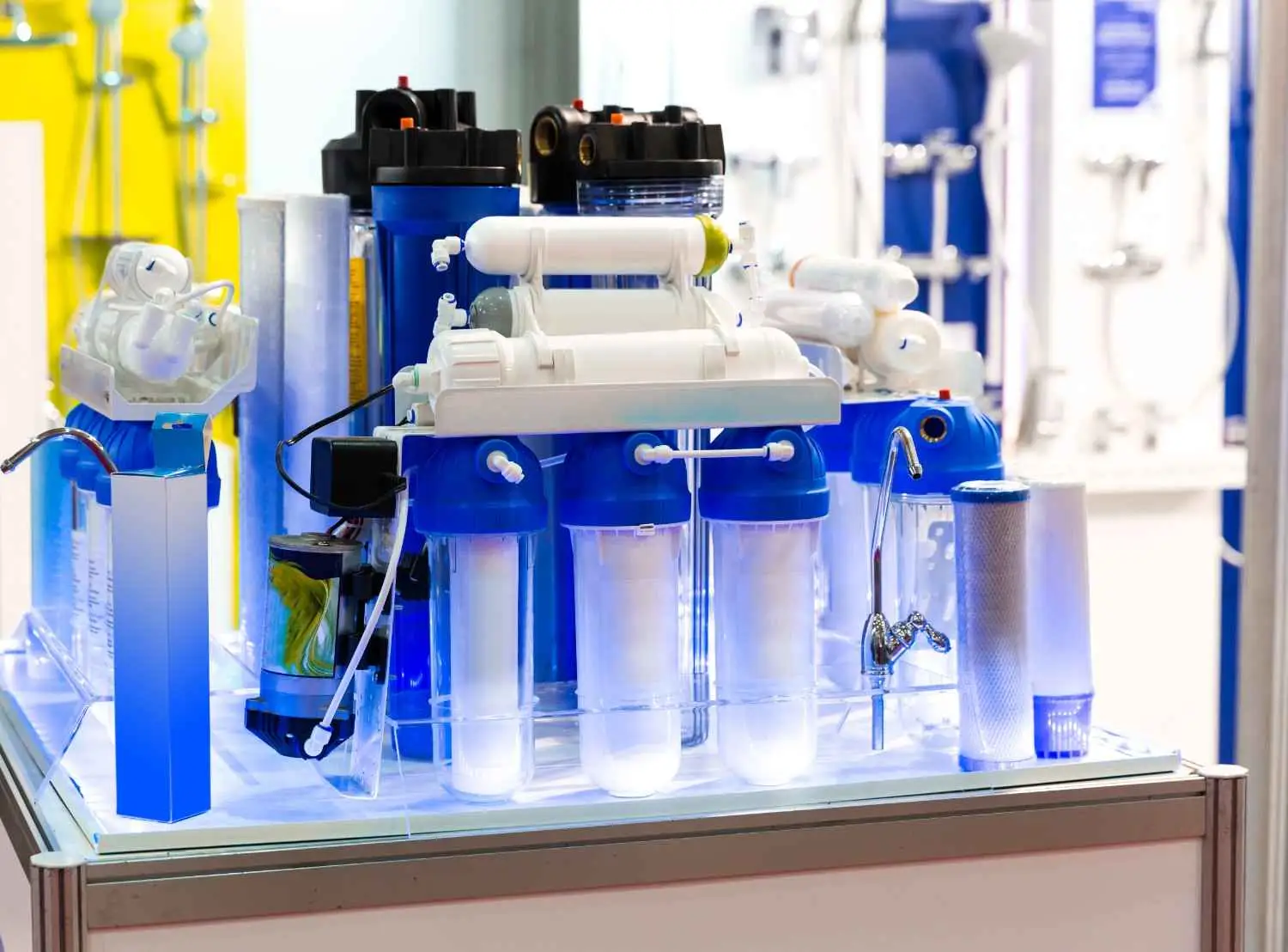 A high-tech reverse osmosis water purification system featuring blue and transparent vertical filter housings, horizontal membrane canisters, and a chrome dispensing faucet on a display stand.