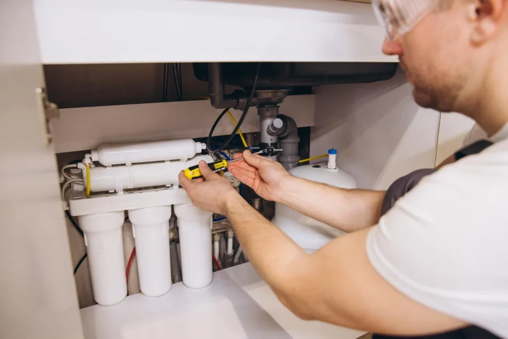Professional plumber installing a multi-stage reverse osmosis (RO) water filtration system under a kitchen sink using a screwdriver.